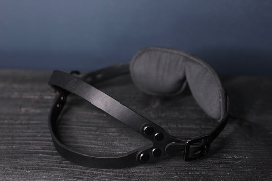 A view of the black leather blindfold from behind, showing the soft jersey eyepad, and the dual strap head harness.
