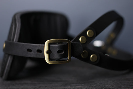 This is a closeup of a bronze buckle on a black leather blindfold sitting on a black surface. The double-strap fit for a secure hold from the head harness portion of the blindfold during bdsm play is visible.