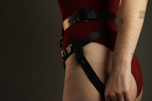An individual is pictured in profile, from the ribs to the upper thighs, wearing a red leather body suit and a black leather strapon harness. The waist band sits at their waist, and below it the hip belt connects to a strap that secures the strapon harness under their buttcheek. 