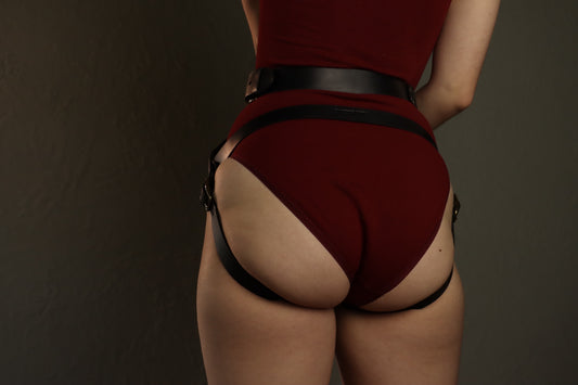 An individual is pictured from behind, from the lower ribs to upper thighs. They are wearing a red bodysuit under a black leather strapon harness. The strapon harness has leather straps securing it at the waist, above the butt, and under each butt cheek.