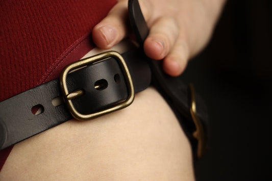A closeup of the brass buckle on a black leather strapon harness, being held by the individual wearing it. 