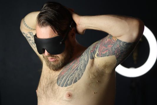 A person with brown hair, a full beard, and several arm tattoos is pictured from the front with their hands behind their head, wearing a black leather blindfold with gunmetal grey fixtures.