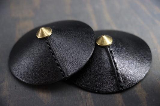 Black leather nipple covers  with brass tips pictured resting on wood. This is a closeup of the seam, which is decorative and functional. 