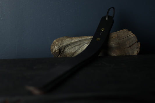 The spanking toy is viewed draped over a piece of driftwood. It is three similarly sized strips of black leather, secured with two rivets at the grip and a leather strap for hanging or carrying. 