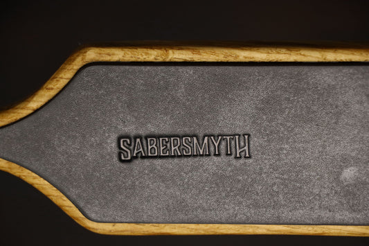 Closeup of the leather inlay on the oak spanking paddle, surrounded by oak wood. The leather insert makes a unique impact play sensation. The brand name, Sabersmyth, is pressed into the leather.