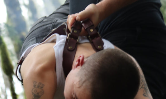 A person wearing the Oliver leather harness is pictured being carried by the straps on the back of the harness. They are positioned face down, the bondage harness is holding their weight in a distributed manner across their torso.