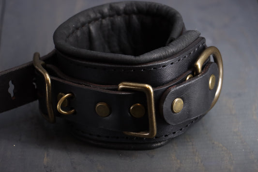 Closeup of a black leather bdsm cuff, lined with deerskin, with bronze fittings. 