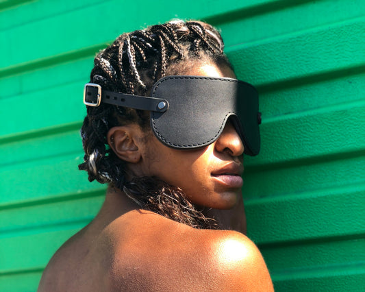 A person with deep brown braids stands against a green wall, head angled over their shoulder. They are wearing a black leather blindfold with black fixings. 