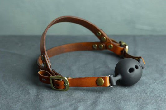 A cognac brown leather head harness ball gag rests on a grey surface. On the left, the two leather straps that cup the wearer's head are visible. On the right, a silicone ball gag that snaps into place. There's a bronze buckle on each side of the ballgag head harness.