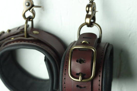 A closeup on a pair of lined leather cuffs with bronze details, hanging against a wall.