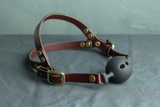 An oxblood red leather ball gag head harness with bronze hardware sits on a grey cloth. A black silicone ballgag is fitted into the harness. The back has two straps coming out from the over-ear buckle at a roughly 45 degree angle, so the harness cradles the wearers head at the base of the skull and higher up.  