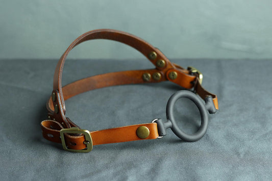 A cognac brown leather head harness with bronze hardware sits on a grey cloth. A black silicone ring gag is fitted into the harness. The back has two straps coming out from the over-ear buckle at a roughly 45 degree angle, so the harness cradles the wearers head at the base of the skull and higher up on the wearer's head.