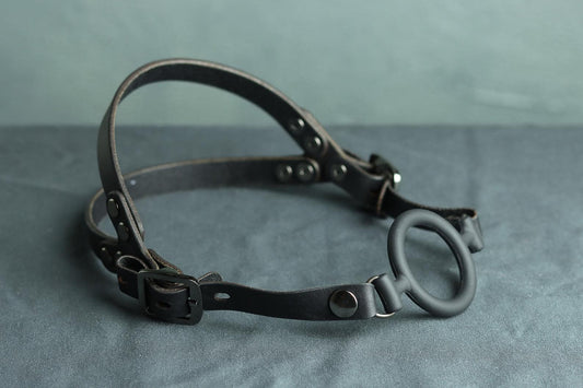 A black leather head harness sits on a grey cloth. A black silicone ring gag is fitted into the harness. The back has two straps coming out from the over-ear buckle at a roughly 45 degree angle, so the leather harness cradles the wearers head at the base of the skull and higher up on the wearer's head.