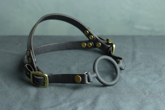 A black leather head harness with bronze hardware sits on a grey cloth. A black silicone ring gag is fitted into the harness. The back has two straps coming out from the over-ear buckle at a roughly 45 degree angle, so the harness cradles the wearers head at the base of the skull and higher up on the wearer's head.