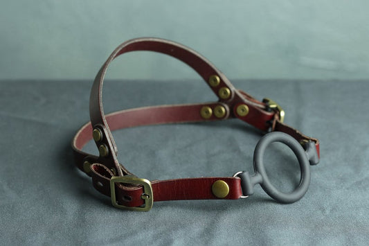 An oxblood red leather head harness with bronze hardware sits on a grey cloth. A black silicone ring gag is fitted into the harness. The back has two straps coming out from the over-ear buckle at a roughly 45 degree angle, so the harness cradles the wearers head at the base of the skull and higher up on the wearer's head.