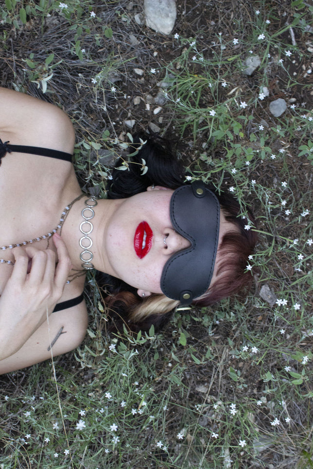 Luxury leather blindfold, worn by woman with red lipstick laying on a grassy field of flowers