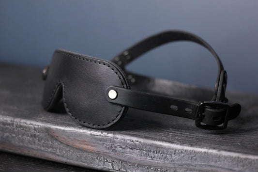 A petite blackleather blindfold with black hardware sits on a black wood surface. The double strapped head harness is visible. 
