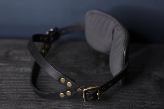 A black leather blindfold sits on a black surface. The double-strap fit for a secure hold during bdsm play is visible. There are bronze fittings and buckles on each side of the blindfold. 