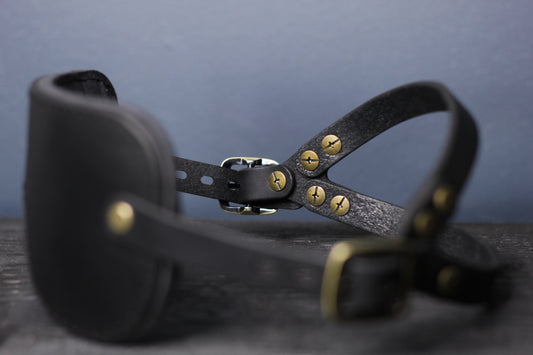 This is a closeup of athe bronze fittings on a black leather blindfold sitting on a black surface. The double-strap fit for a secure hold during bdsm play is visible.