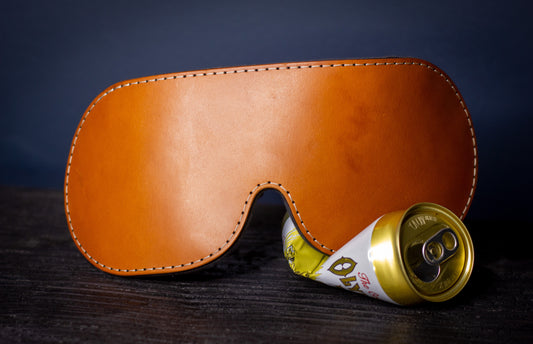 A cognac brown leather blindfold with a weighted flaxseed back sits on a black piece of wood.  The stitching is tan. The blindfold is sitting on a crushed yellow can,.
