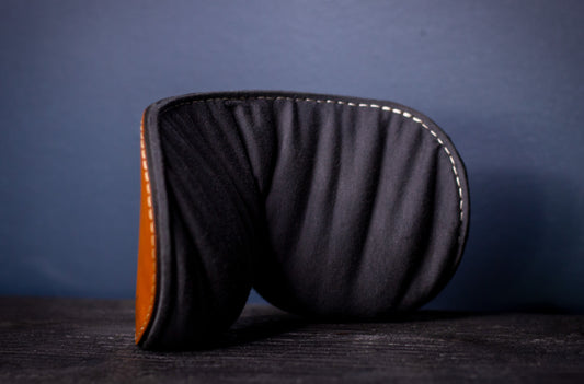 A cognac brown leather blindfold with a weighted flaxseed back sits on a black piece of wood. This image shows the leather blindfold from behind, where a soft jersey fabric  heavy with flaxseed invites the wearer to relax.