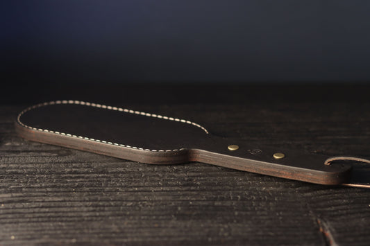 The black leather paddle is pictured resting on a dark wood surface. It has neat stitching around the edges and a looped piece of leather on the handle for storage. 