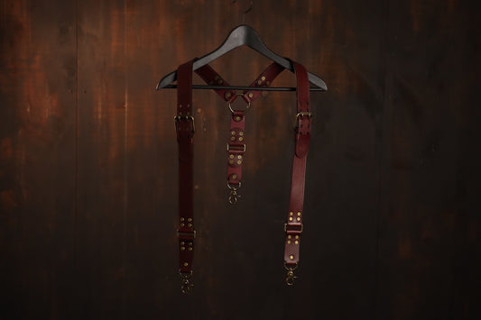 The suspenders are pictured hanging on a coat hanger. The suspenders feature a beautiful and functional buckle that rests near the shoulders, brass clips at the bottom, and brass rivets. The back has an o ring where the two straps meet and a single suspender strap connects to the back of the wearer's pants or leather harness. 