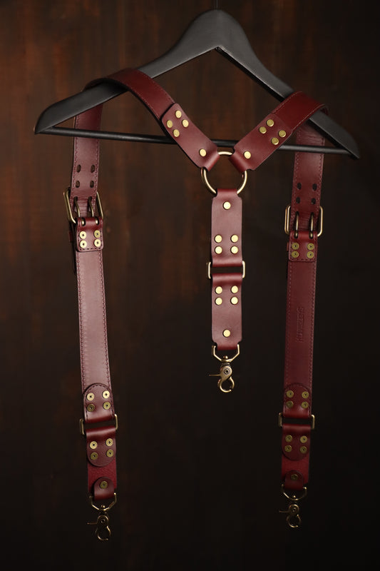 The suspenders are pictured hanging on a coat hanger. The suspenders feature a beautiful and functional buckle that rests near the shoulders, brass clips at the bottom, and brass rivets. The back has an o ring where the two straps meet and a single suspender strap connects to the back of the wearer's pants or leather harness. 