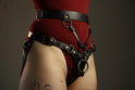An individual is pictured from the lower ribs to upper thighs, wearing a red bodysuit under a black leather strapon harness. The leather strapon harness has a waist belt, a front plate, straps at the hip, and stabilizing sraps connecting the waist belt to the front plate. 