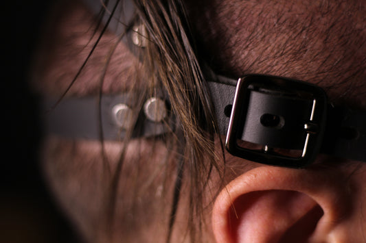 A closeup of a black buckle on the head harness over a person's ear. On the side, the way the head harness of this blindfold cups their head is visible. 