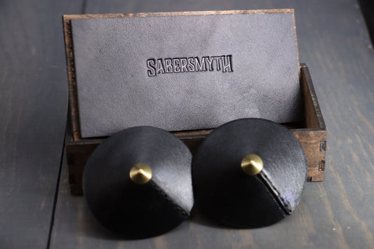 Black leather nipple covers pictured resting against a wood box. They have small triangular brass rivets at the center.  The wooden box is inlayed with leather on the lid, with the name Sabersmyth pressed into the leather. 