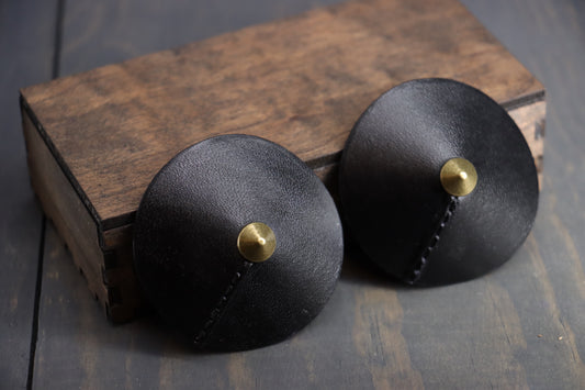 Black leather nipple covers pictured resting against a wood box. They have small triangular brass rivets at the center.  