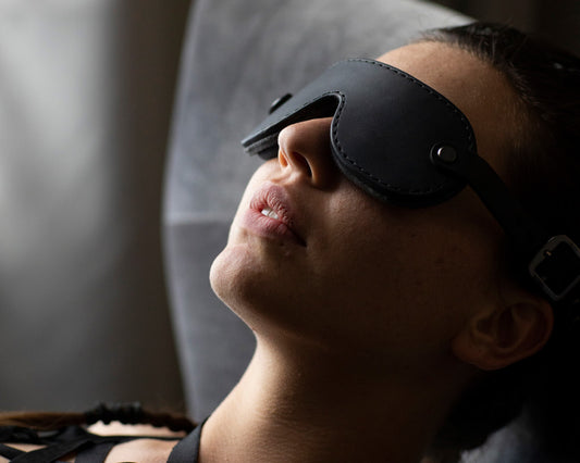 A person wearing a small black leather blindfold is viewed reclining in natural light peacefully.