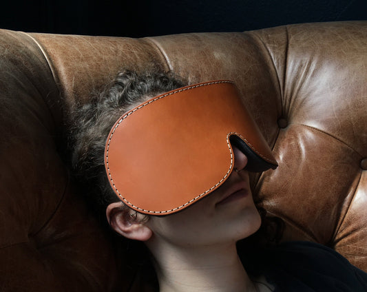 An individual with curly brown hair and a peaceful smile rests wearing the weighted eye pillow on a brown leather sofa. The blindfold is brown, and the size of roughly half their face, with the top edge resting on their hairline, and the bottom edge resting just below their cheekbones