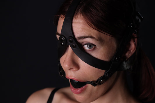 A person wearing the the locking leather head harness looks at the camera. Two straps join at the bottom to hold the ballgag in place, criss crossing over the cheeks with straps that join between the eyebrows and connect to the back of the head harness.