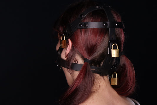 A view of the head harness form the back, on a person with red pigtails. One strap reaches underneath the ears and buckles at the base of the neck. Another head harness strap connects from over the top of the head. Each bdsm head harness strap visible is adjustable. Three locks are visible from this angle, one over the ear and two along the back of the wearer's head, securing the locking head harness. 