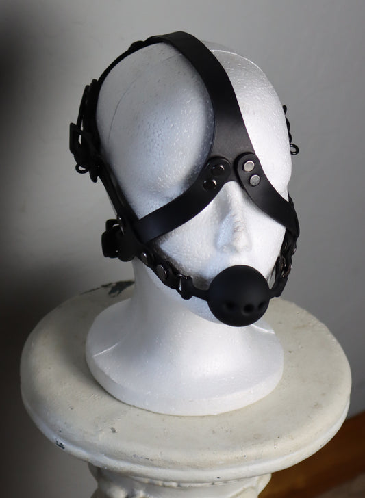 The locking leather head harness is displayed on a mannequin head. Two straps join at the bottom to hold the ballgag in place, criss crossing over the cheeks with straps that join between the eyebrows and connect to the back of the head harness.