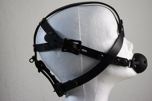 The locking leather head harness is displayed on a mannequin head in profile. Two straps criss cross across the cheek, with one connecting to the ballgag, and another crossing under the eye up to between the eyebrows, where it arches over the top of the head. There are several straps on the back of this locking head harness, with multiple adjustment and lock opportunities for a secure fit during a bondage scene. 