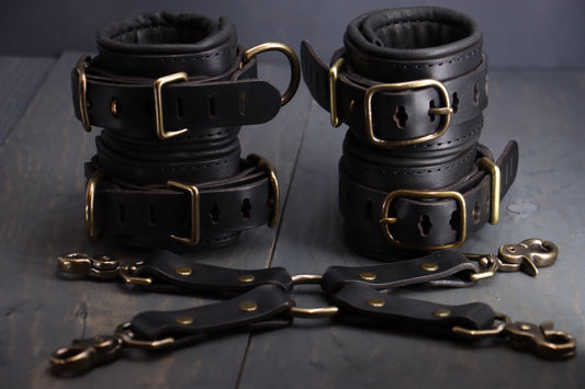 Four black leather cuffs with bronze fittings sit on a grey wood surface, with a 4 way leather attachment alongside them.