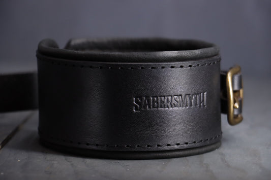 Closeup of the sabersmyth logo pressed into a black leather bdsm cuff, with bronze fittings and black deerskin lining.