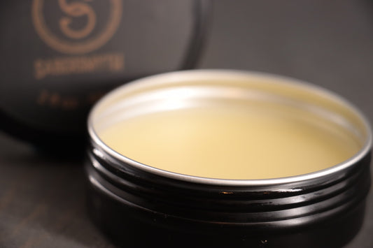a jar of leather conditioner sits open on a wood surface. The leather conditioner is a blend of oils designed to keep BDSM gear looking and feeling supple and flexible. 