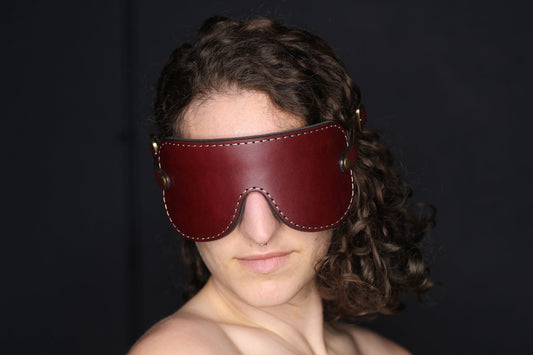 A person with brown curly hair is pictured from the shoulders up wearing an oxblood red leather blindfold with bronze fittings. 