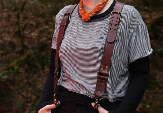 An individual is wearing the leather suspenders over a striped black tee shirt, with black pants and an orange scarf.  The suspenders feature a beautiful and functional buckle that rests near the shoulders, brass clips at the bottom, and brass rivets. 