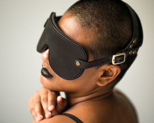 A person with short cropped hair and black lipstick is viewed from the back, leaning their head over their shoulder, wearing a black leather blindfold with bronze fittings. The buckle is visible over their ear, and the head harness portion of the blindfold cradles the back of their head.