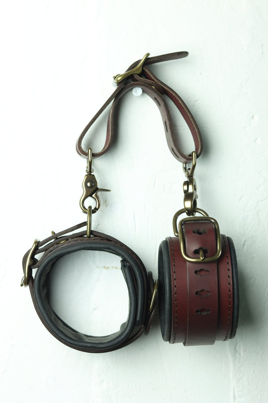 A pair of lined bdsm leather cuffs hangs on a wall, connected with the attachment belt for flexibility in use. 