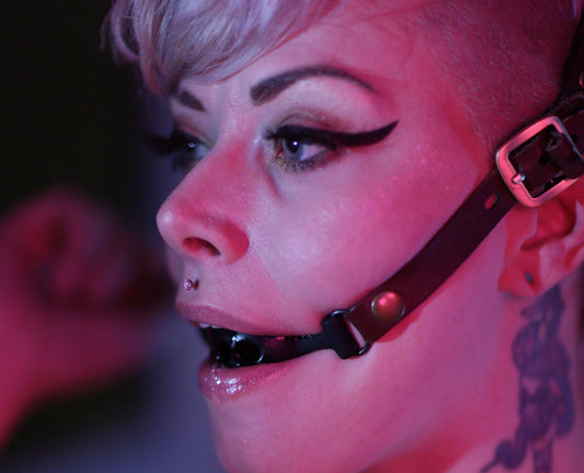 A blonde person with a neck tattoo, cat-eye makeup, and a medusa or philtrum piercing is photographed in profile, wearing a ring gag fitted into a black leather harness. The buckle of the bondage leather ball gag is visible over their ear, the photo is cast in a soft reddish pink light.