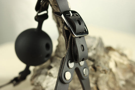 A closeup of the gunmetal grey hardware of this black leather head harness draped over a piece of wood, showing in the foreground the gunmetal grey buckle, and in the background a black silicone ball gag. 