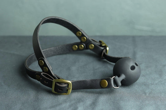 A black leather ball gag head harness with bronze hardware sits on a grey cloth. A black silicone ballgag is fitted into the harness. The back has two straps coming out from the over-ear buckle at a roughly 45 degree angle, so the harness cradles the wearers head at the base of the skull and higher up.