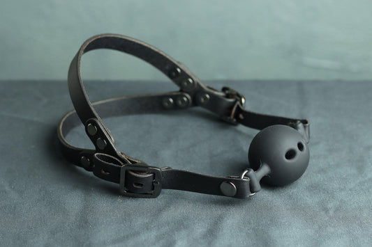 A black leather ball gag head harness with gunmetal grey hardware sits on a grey cloth. A black silicone ballgag is fitted into the harness. The back has two straps coming out from the over-ear buckle at a roughly 45 degree angle, so the harness cradles the wearers head at the base of the skull and higher on the head.