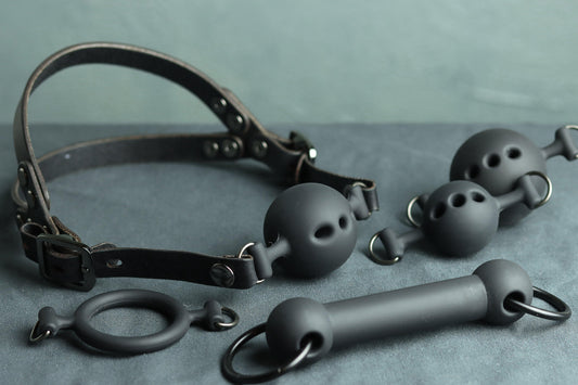A black leather ball gag head harness sits on a grey cloth. A black silicone ballgag is fitted into the harness, next to it are two additional ball gags in additional sizes (a large ball gag, and a small ball gag). Underneath, there is a silicone ring gag and a silicone bit gag. The back has two straps coming out from the over-ear buckle at a roughly 45 degree angle, so the harness cradles the wearers head at the base of the skull.